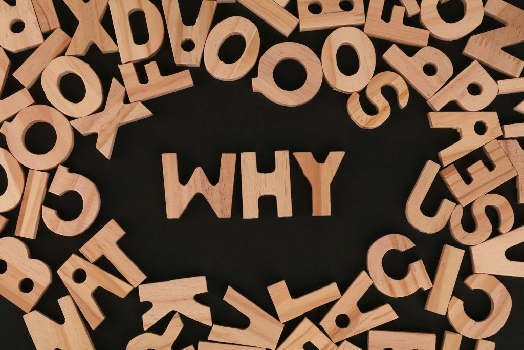 Scattered wooden alphabet letters with the word WHY at the center on a black background in a flat lay arrangement.