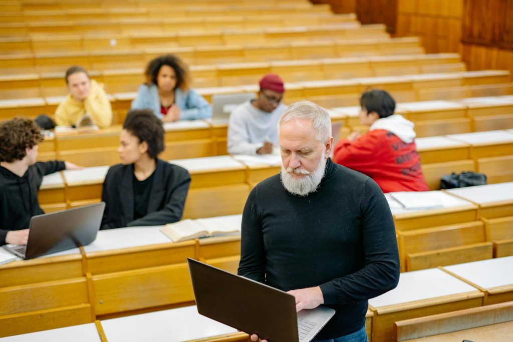 CHED Tertiary Education Subsidy Solves Financial Qualms to Conquer Dream Career 6 Mature teacher interacting with diverse students using laptops in a spacious lecture hall setting.
